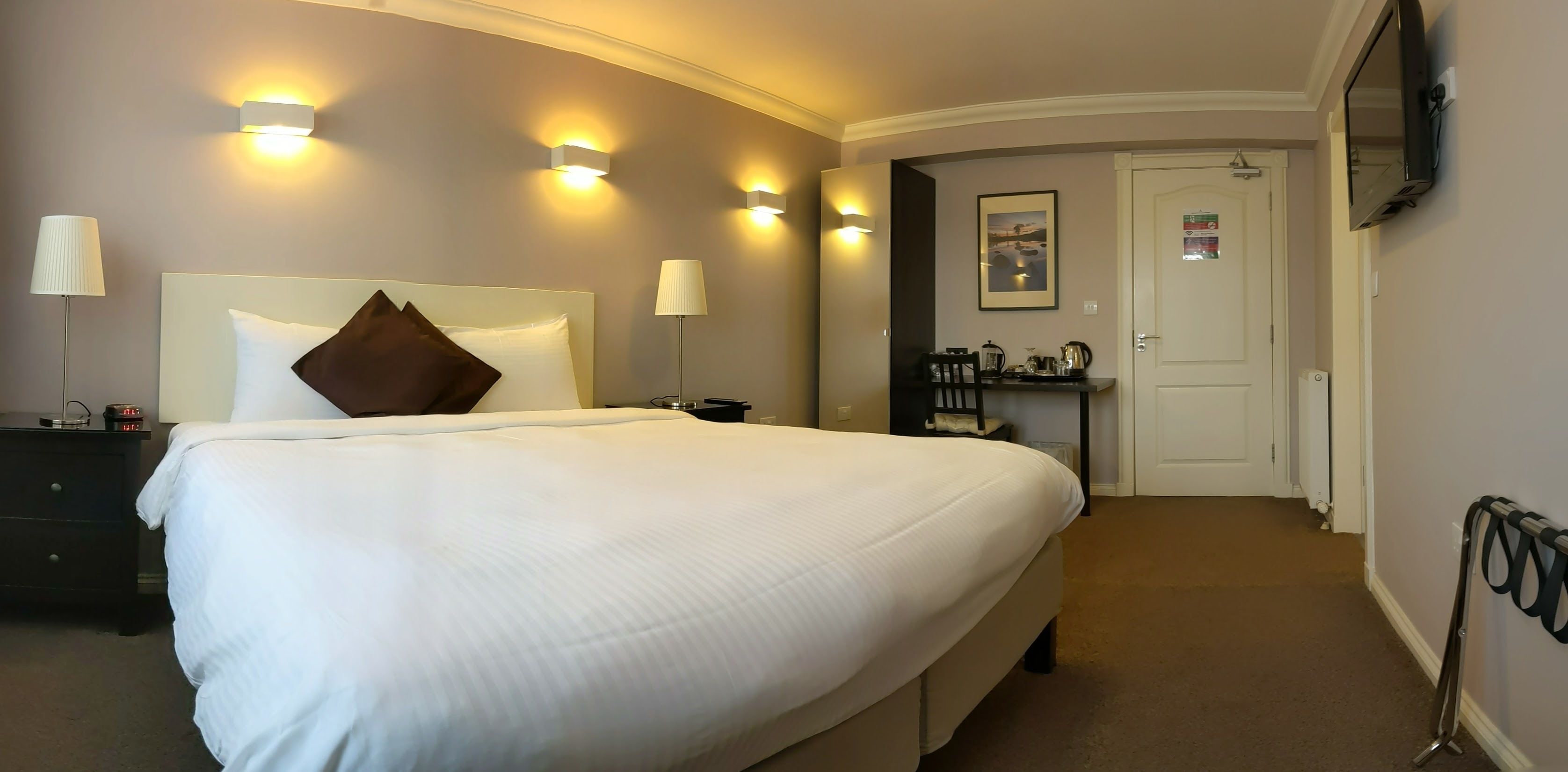 A hotel room with a large bed, bedside lamps, a desk with a chair, a television, and a door leading to another room.