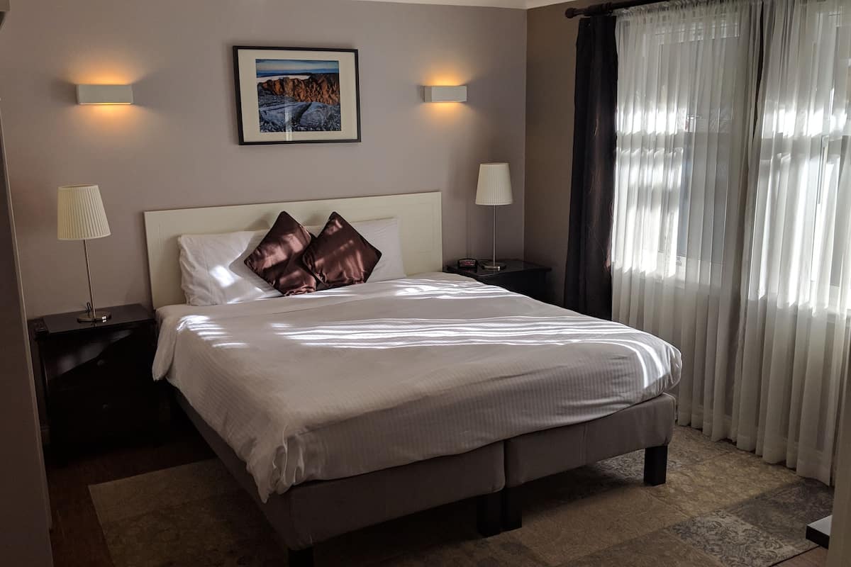 A neatly made bed with pillows, a bedside lamp, a framed picture on the wall, and a window with curtains.