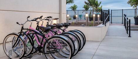 A row of bicycles parked against a building wall, with a view of the ocean and palm trees in the background.