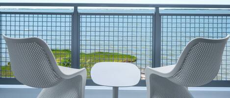 A balcony with two white chairs and a small table, overlooking a body of water and a distant shoreline.