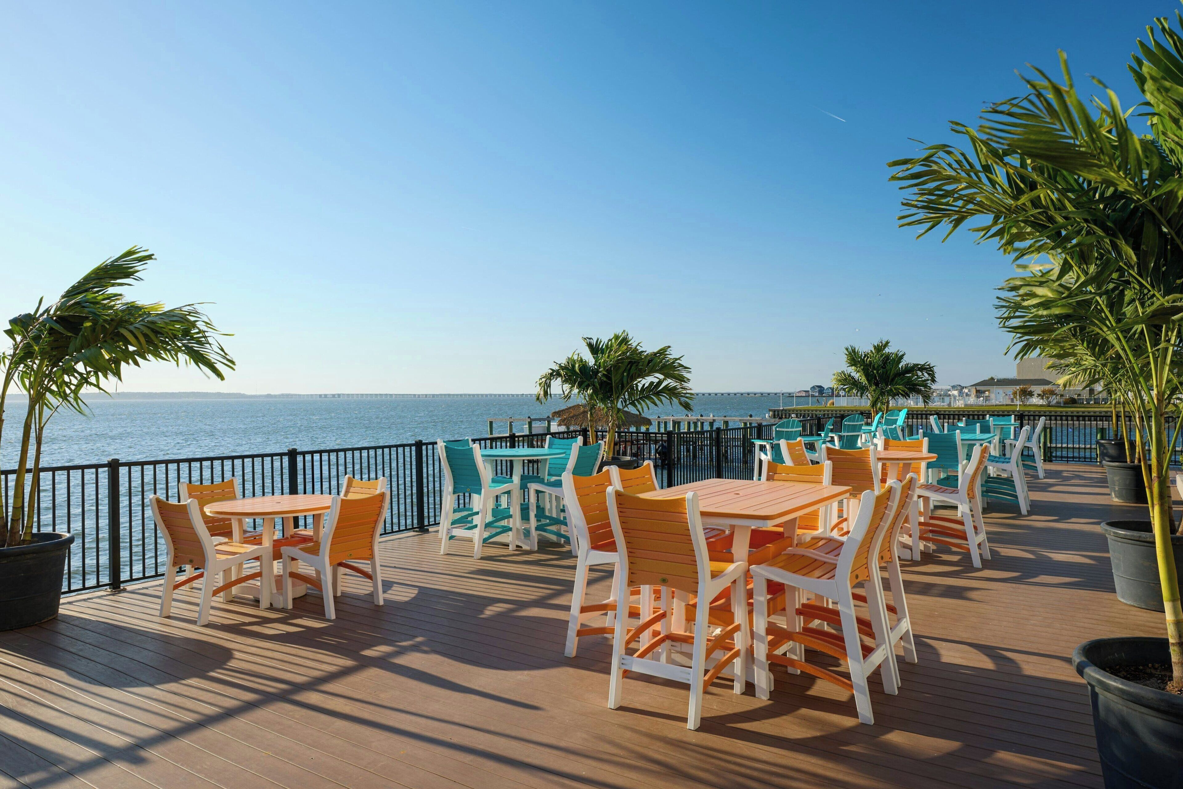 A deck with tables and chairs, overlooking a body of water, with a railing and potted plants.