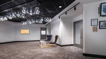 Un vestíbulo amplio con diseño moderno, que incluye una gran zona de estar con sillones y una mesa, una alfombra estampada y un techo con lámparas colgantes.
