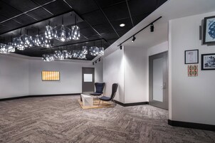 A spacious lobby with a modern design, featuring a large seating area with chairs and a table, a patterned carpet, and a ceiling with hanging lights.