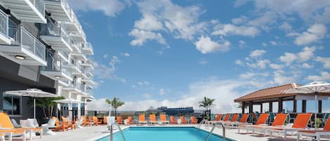 Ein Swimmingpool mit orangefarbenen und weißen Liegestühlen, einem klaren, blauen Himmel und einem modernen Gebäude mit Balkonen.
