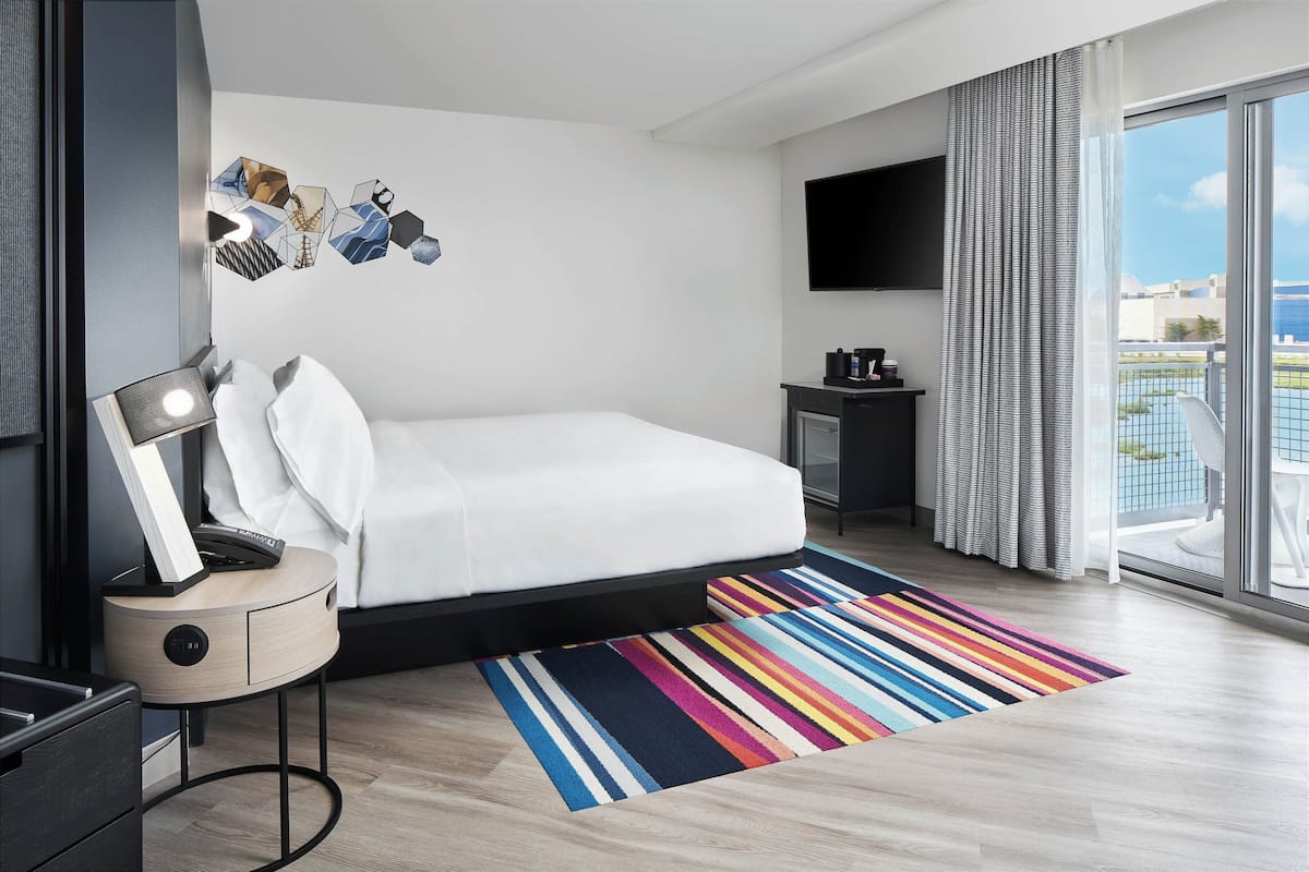A modern hotel room with a bed, a bedside table with a lamp, a flat-screen TV, a small side table with a coffee maker, and a large window with a view of the city.