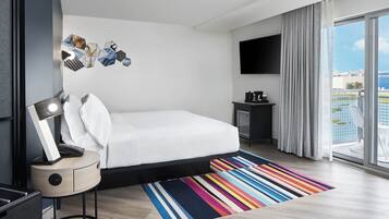 A modern hotel room with a bed, a bedside table with a lamp, a flat-screen TV, a small side table with a coffee maker, and a large window with a view of the city.