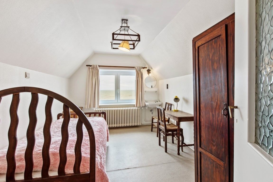 A bedroom with a sloped ceiling, a wooden bed, a desk with a chair, a window with curtains, and a door.