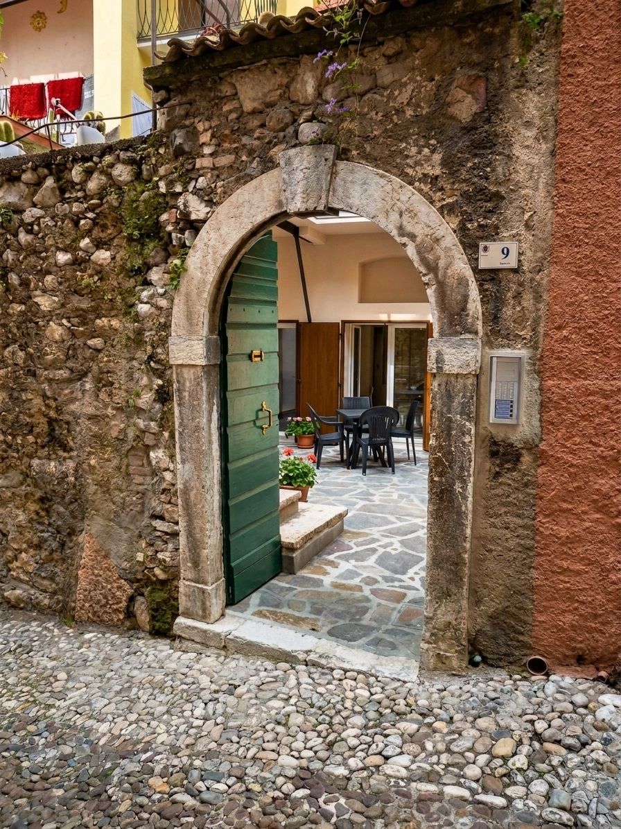 An arched stone doorway leading to a courtyard with a stone floor, a green door, and a small outdoor seating area with tables and chairs.