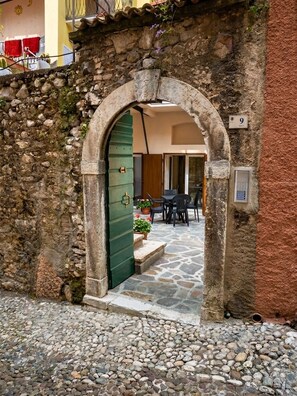 Un arcoado acceso de piedra que da a un patio con suelo de piedra, una puerta verde y una pequeña zona de estar al aire libre con mesas y sillas.