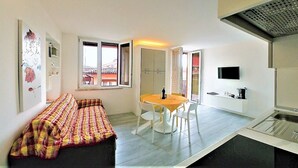 Un espacio habitable compacto con sofá, una mesa de comedor con sillas, una zona de cocina con fregadero y un televisor montado en la pared.