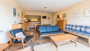 A cozy living room with a sofa, chairs, and a wooden table. There is a kitchen area in the background. - Zandstuyver 102 (Koksijde)