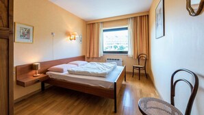 A hotel room with a wooden bed, a nightstand with a lamp, a chair, a window with curtains, and a framed picture on the wall. - Zandstuyver 102 (Koksijde)