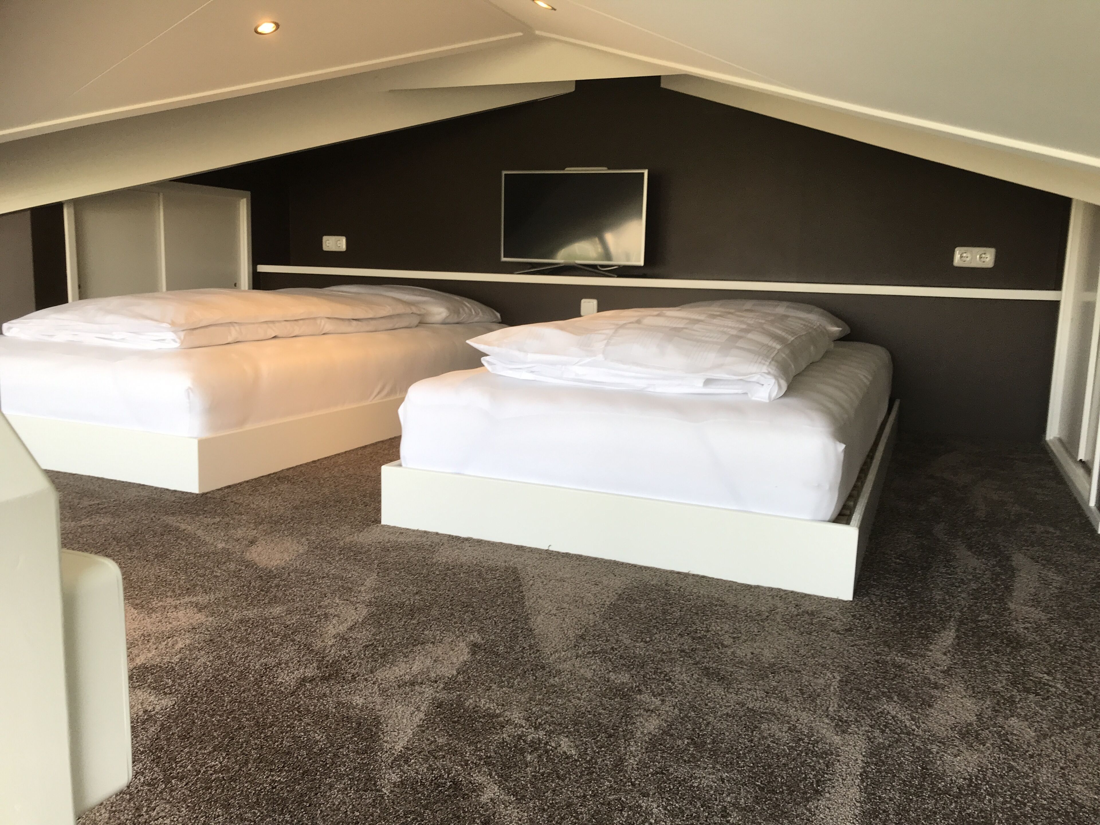A small, modern room with two white beds, a flat-screen TV mounted on the wall, and a carpeted floor.