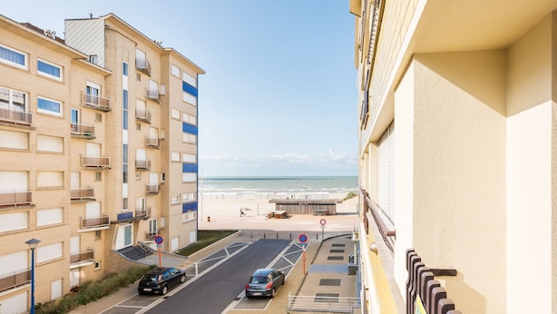 Eine Küstenstraße mit Blick auf den Strand, ein geparktes Auto am Straßenrand und ein Gebäude mit Balkonen.