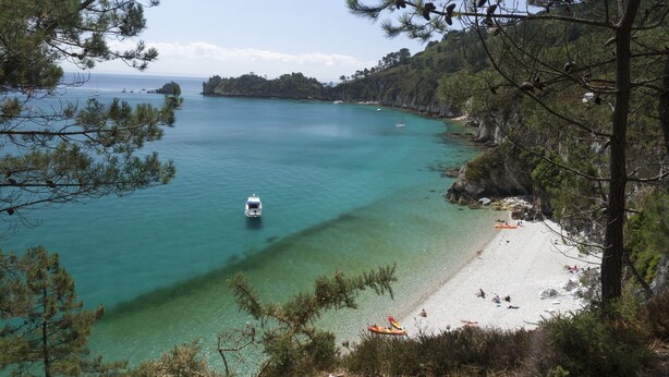 Una platja costanera amb aigües turqueses cristal·lines, un vaixell petit, i una riba sorrenca amb gent i un caiac.