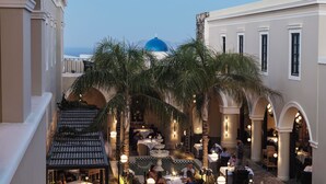 A courtyard with tables and chairs, palm trees, and a blue dome in the background. - Katikies Garden Santorini - The Leading Hotels Of The World (Santorini)