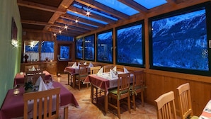 Zona pranzo con tavoli e sedie di legno, ampie finestre con vista sulle montagne e soffitto in legno con lucernari.