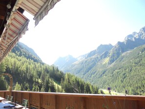 A wooden deck with chairs, overlooking a mountainous landscape with dense forests and peaks.