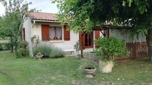 Une maison avec un toit en tuiles, une clôture en bois et un jardin agrémenté de plantes en pot et d’un banc.