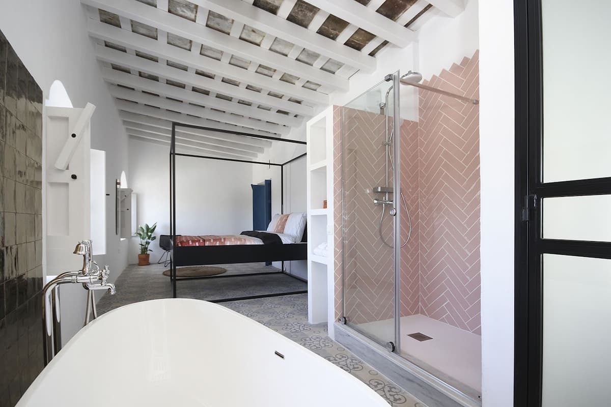 A modern bathroom with a freestanding bathtub, a glass-enclosed shower, and a bed in the background.