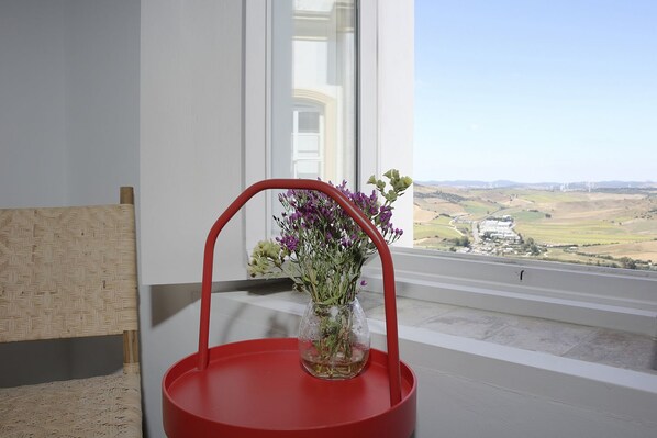 Una silla roja con un florero de vidrio que contiene flores moradas, ubicada junto a una ventana con vistas a un paisaje rural.