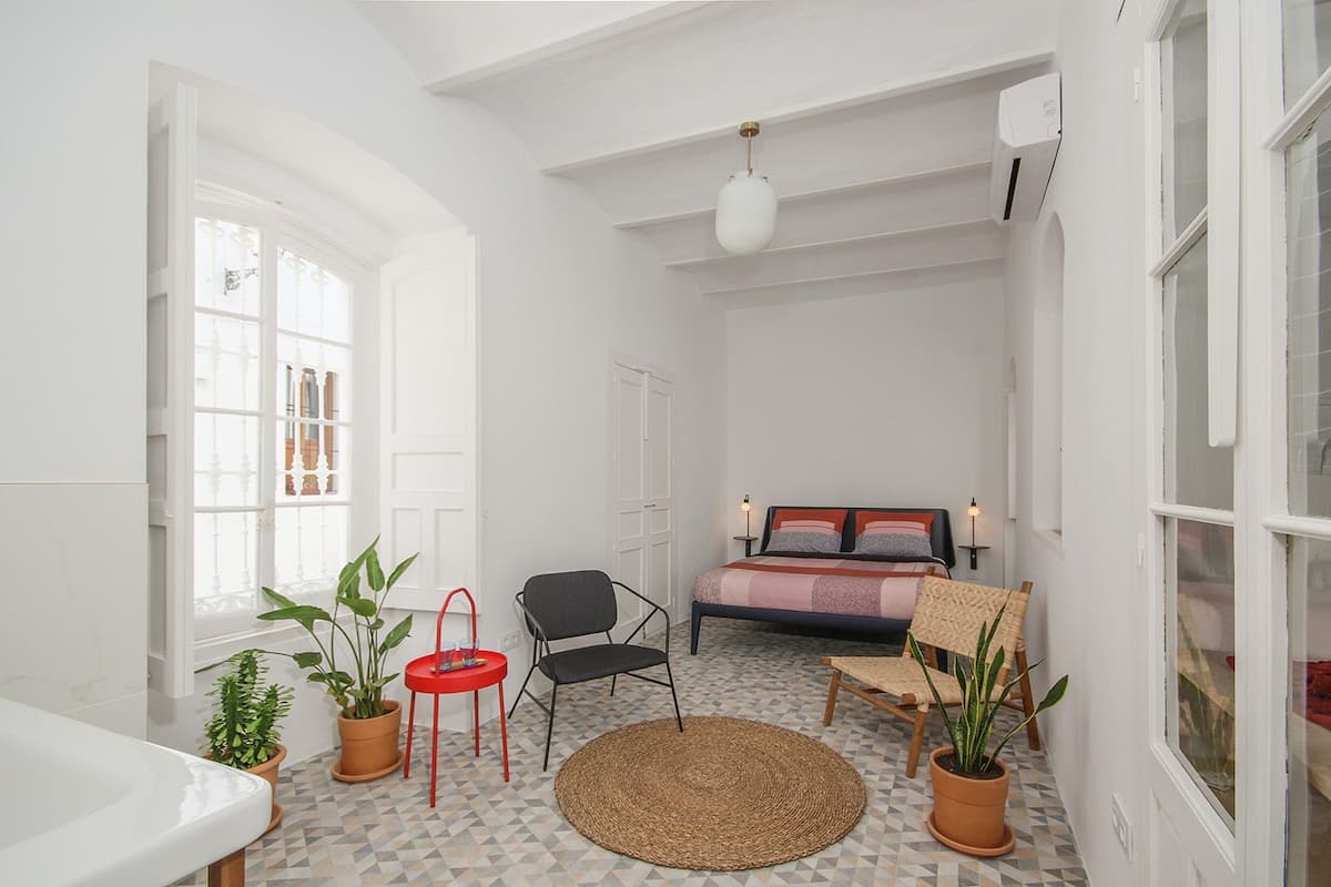 A compact bedroom with a bed, two chairs, a small table, and potted plants.