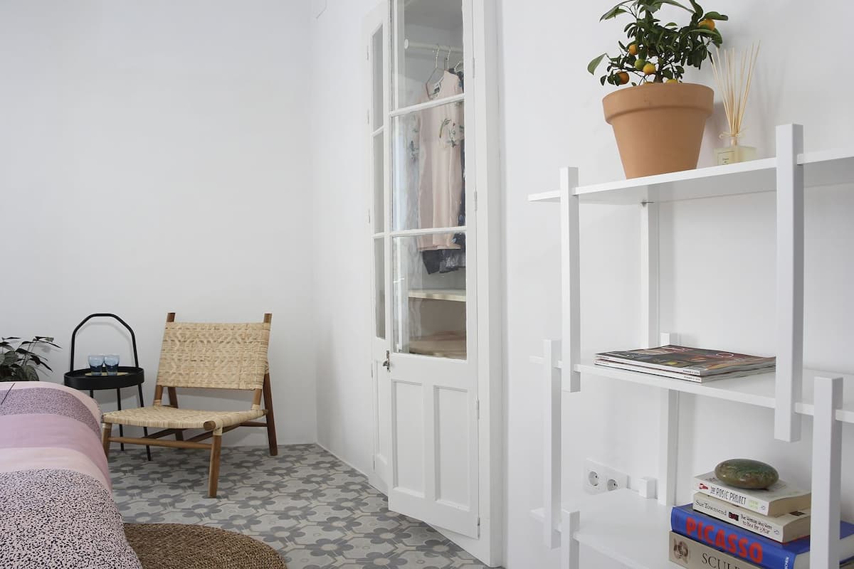 A room with a white wardrobe, a wicker chair, a potted plant, and a bookshelf with books.