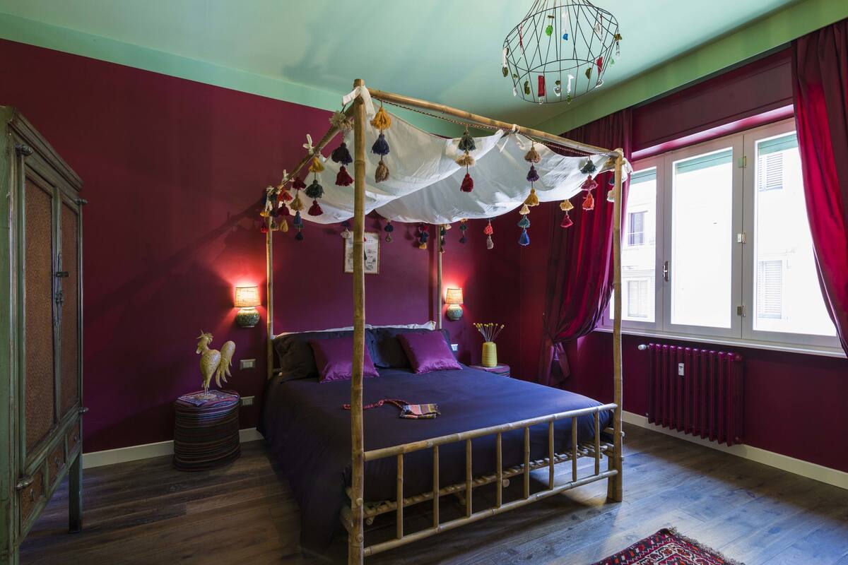 A four-poster bed with a canopy, a red wall, a window with curtains, and a radiator.