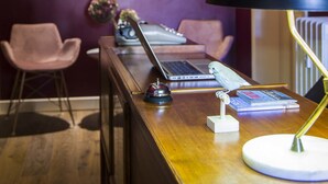 Un espace d’accueil avec un bureau en bois, une lampe noire, un ordinateur portable et une statuette d’oiseau. On y trouve deux chaises roses et un miroir accroché au mur.