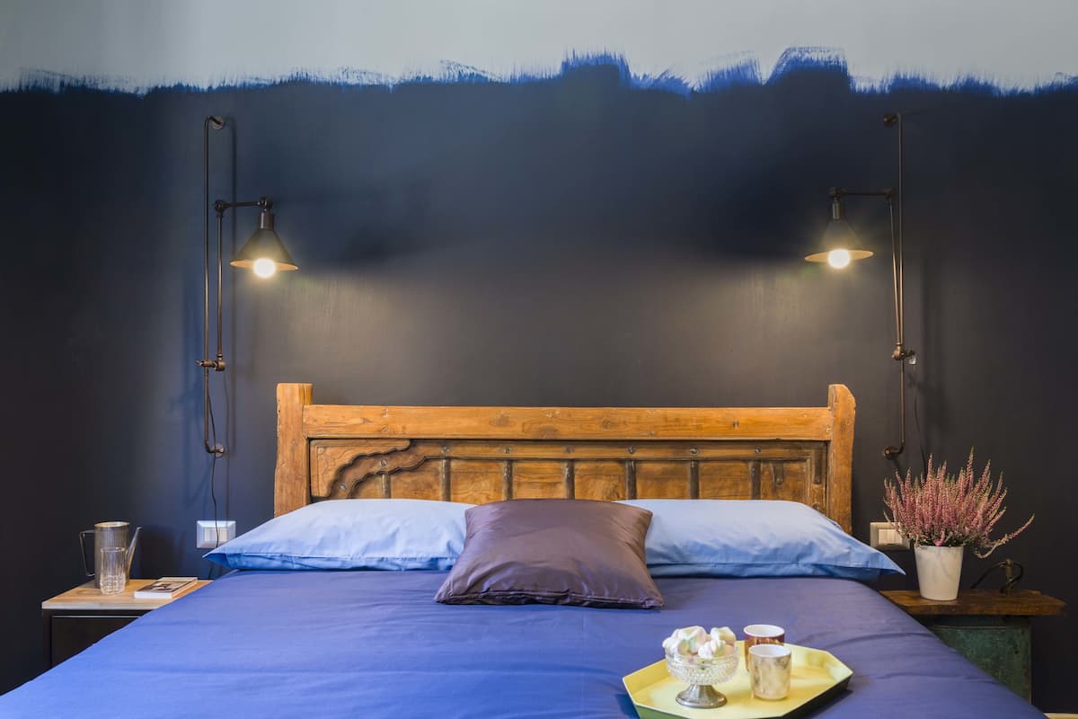 A neatly made bed with a wooden headboard, two bedside lamps, a tray with a teapot and cups, and a potted plant on a nightstand.