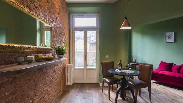 Une pièce avec un mur en briques, un grand miroir, une table à manger avec des chaises, un canapé rouge et une fenêtre donnant sur des immeubles.