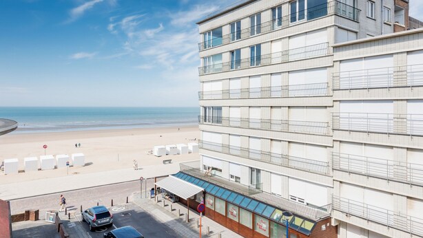 Ett hotell med flera våningar och utsikt över stranden, ett takförsett utomhusområde med sittplatser och en parkeringsplats med bilar.