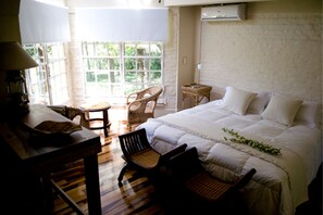 A bedroom with a large bed, a wooden desk, a chair, and a small table. There is a window with a view of greenery outside.