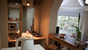 Een keuken met een witte waterkoker, een houten tafel en stoelen. Er is een raam met uitzicht op groen.