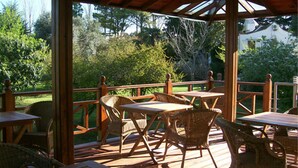 Een terras buiten met houten tafels en rieten stoelen, omringd door een houten terras en met uitzicht op een tuin.