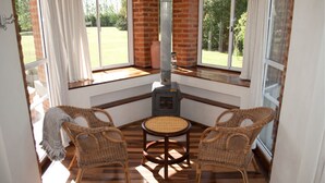 Een hoekzaal met een open haard, rieten stoelen en grote ramen met uitzicht op een tuin.