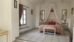 Une chambre à coucher comprenant un lit, une table de chevet, une commode, une fenêtre avec des rideaux, un radiateur et un escalier menant à un autre étage.