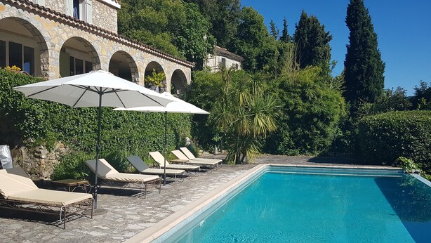 Un espace piscine agrémenté de chaises longues et de parasols, entouré de verdure et d’éléments architecturaux en pierre.