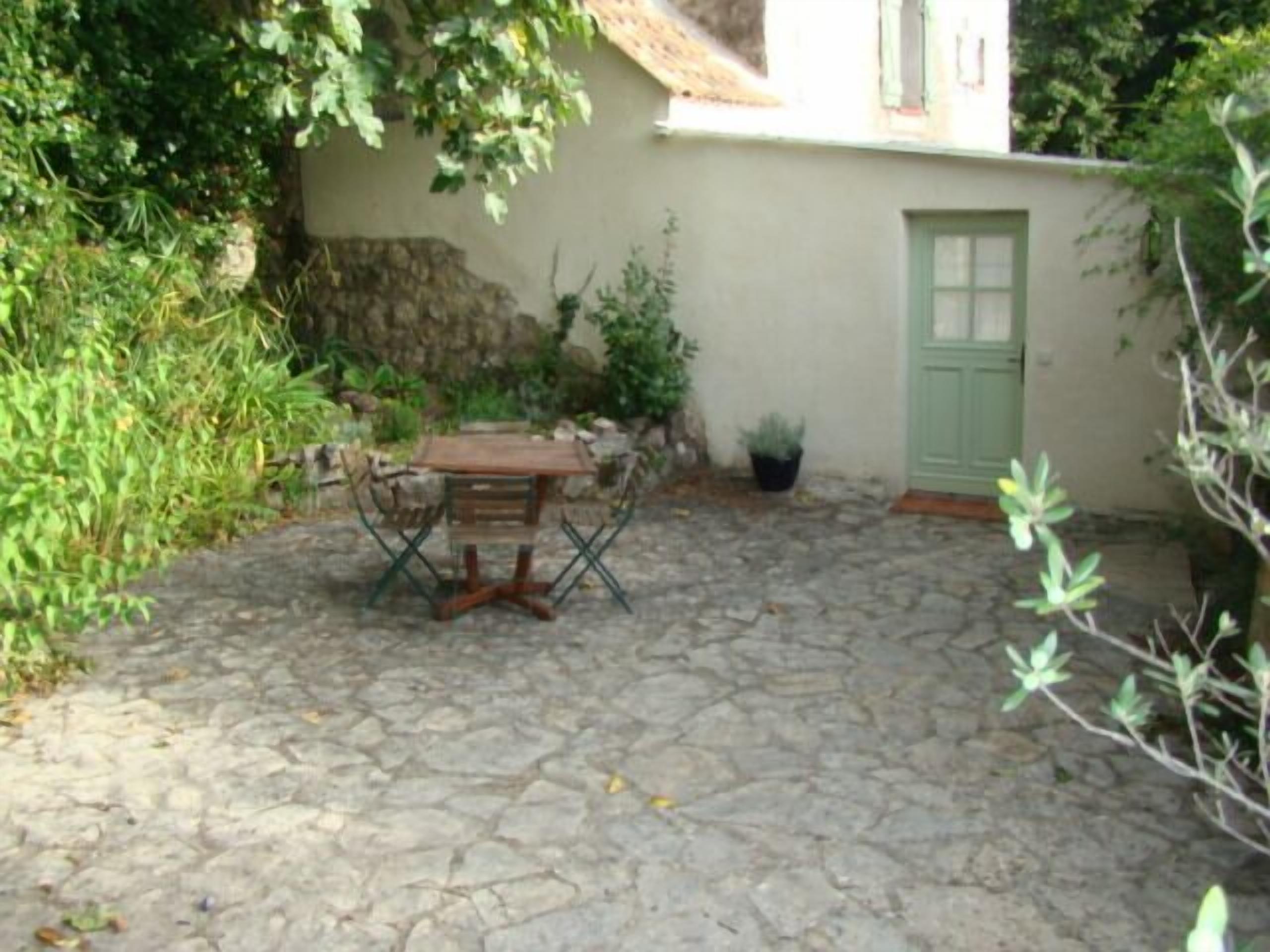 Une terrasse en pierre avec une table et des chaises en bois, entourée de verdure et bordée d’un bâtiment doté d’une porte verte.