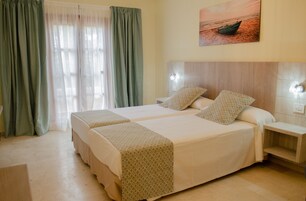 Habitación de hotel con dos camas, una mesita de noche, un cuadro de un barco en la pared y cortinas translúcidas.