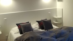 A bedroom with a bed, two bedside tables, a wall-mounted shelf, and a ceiling light fixture.