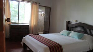 A bedroom with a wooden headboard, a bed with white bedding and a colorful striped blanket, a wooden dresser, and a window with curtains.