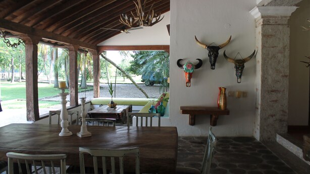 A covered dining area with a wooden table and chairs, a bench, and a view of a garden.