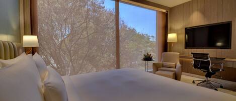 Habitación de hotel con una cama grande, un escritorio, una silla, un televisor y una ventana con vistas a árboles.