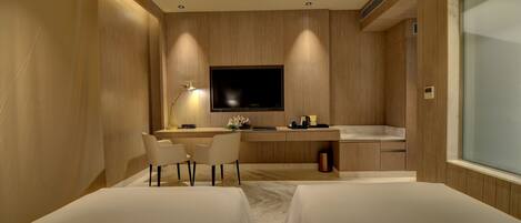 A modern hotel room with a wooden panel wall, a flat-screen TV mounted above a desk, two single beds, a small sitting area with a chair and desk, and a large window.