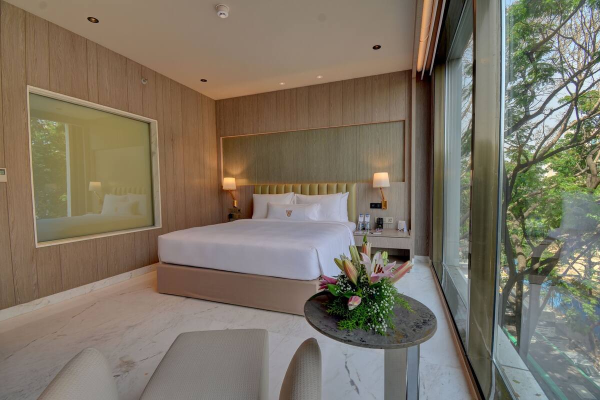 Habitación de hotel moderna con una cama grande, lámparas de noche, una mesita con flores y un ventanal con vistas a árboles.