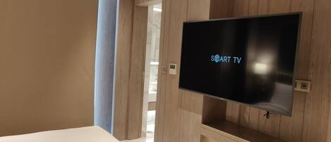 A modern hotel room with a flat-screen TV mounted on a wooden wall, a bed, and a bathroom visible through an open door.