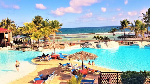 A pool area with lounge chairs and umbrellas, a beach in the background, and palm trees. - Touba Manga Beach (Saint-François)