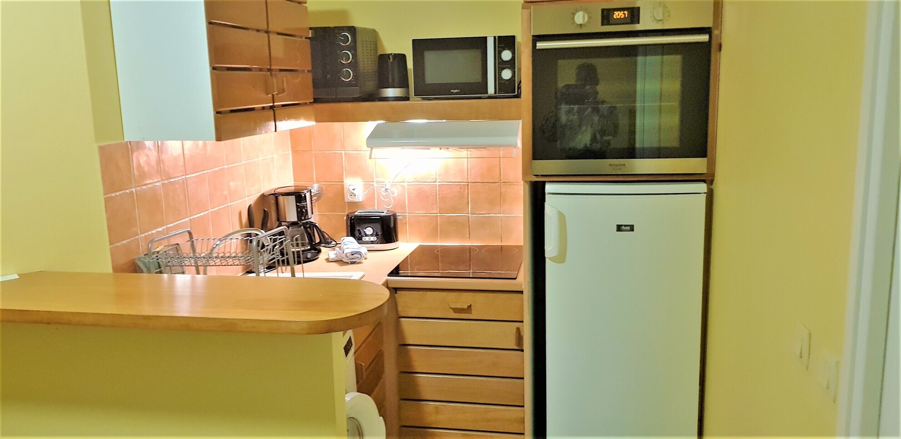 A compact kitchen with a microwave, toaster, and coffee maker on the countertop, a small refrigerator, and wooden cabinets.