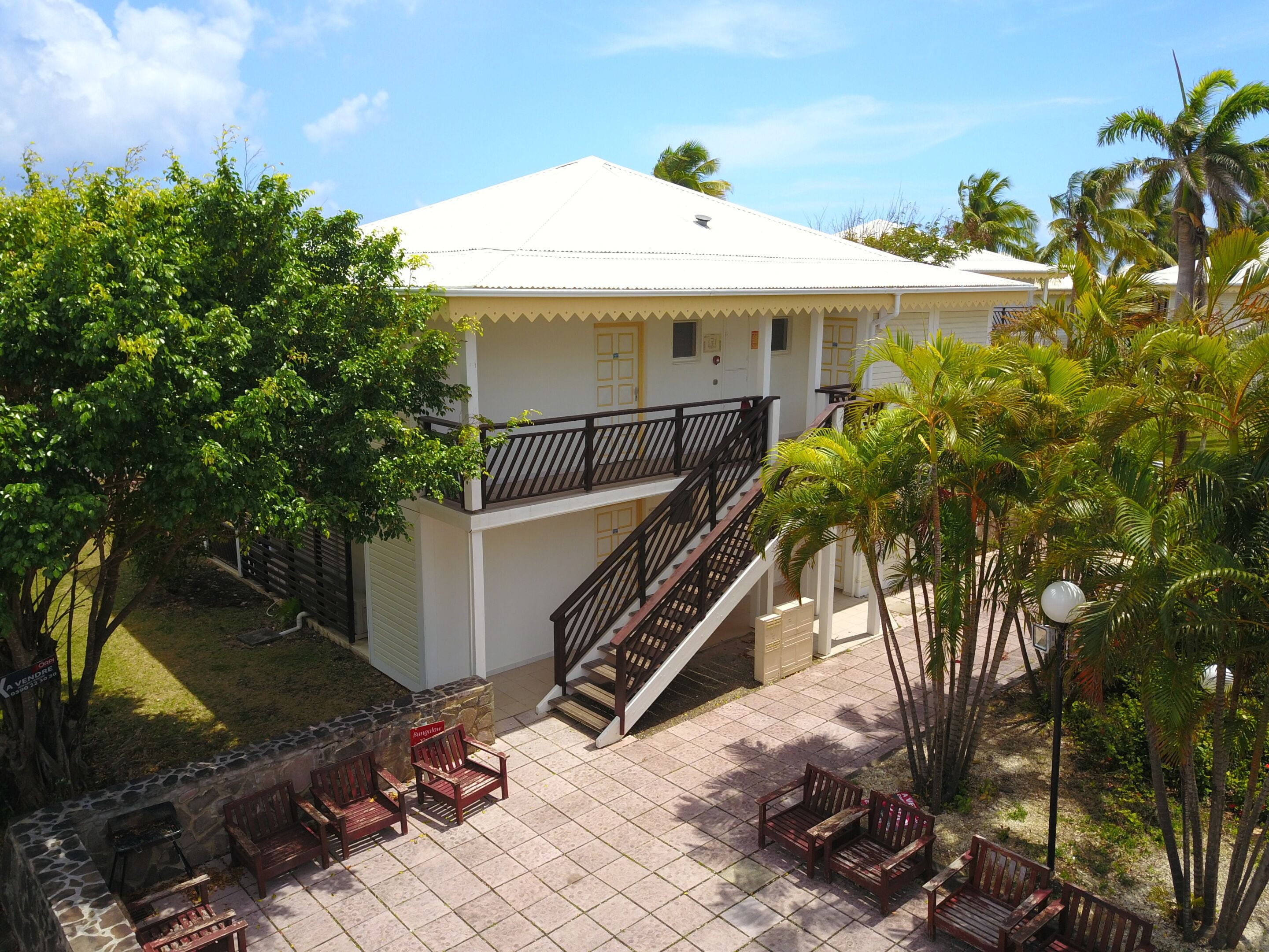 A two-story building with a white exterior, a balcony, and a staircase leading to the upper floor. There are outdoor seating areas with wooden benches and a barbecue grill.